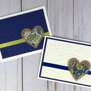 May include: Two handmade greeting cards. One card is navy blue with an embossed pattern, a yellow ribbon, and a burlap heart with a paisley design. The other card is cream with a blue ribbon and a similar heart.