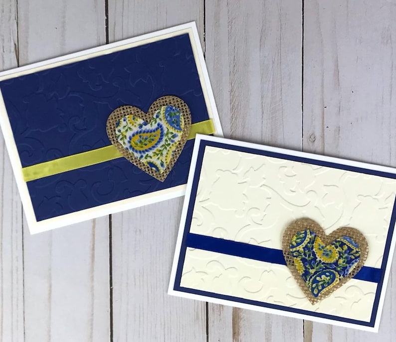 May include: Two handmade greeting cards. One card is navy blue with an embossed pattern, a yellow ribbon, and a burlap heart with a paisley design. The other card is cream with a blue ribbon and a similar heart.