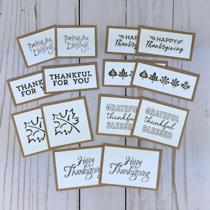 May include: A collection of rectangular paper cards with brown borders and white centers, featuring black ink stamps of autumn leaves and text including Thanksgiving Blessings, Happy Thanksgiving, Thankful For You, and Grateful Thankful Blessed.