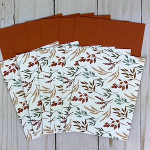 May include: A set of rectangular cards featuring a white background with a repeating pattern of leaves and wheat stalks in shades of rust orange, sage green, and brown. Each card has a solid rust orange back side, fanned out in a stack.