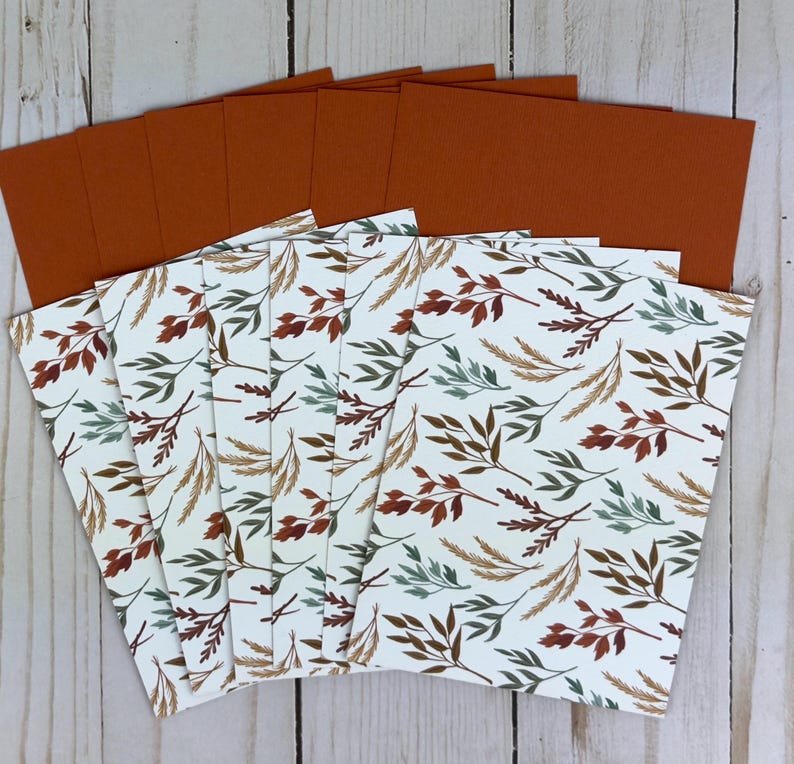 May include: A set of rectangular cards featuring a white background with a repeating pattern of leaves and wheat stalks in shades of rust orange, sage green, and brown. Each card has a solid rust orange back side, fanned out in a stack.