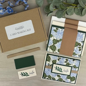 May include: A card making kit in a brown cardboard box labeled E.E. James Stationery. The kit includes envelopes, cardstock, a brown ribbon, a small dark green rectangle, and a white rectangular tag with a leaf illustration and the word hello.