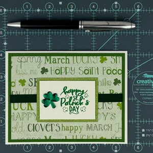 May include: A St. Patrick's Day greeting card with a green and white background featuring the words "Happy St. Patrick's Day" and shamrock designs. A black and silver pen is placed above the card.