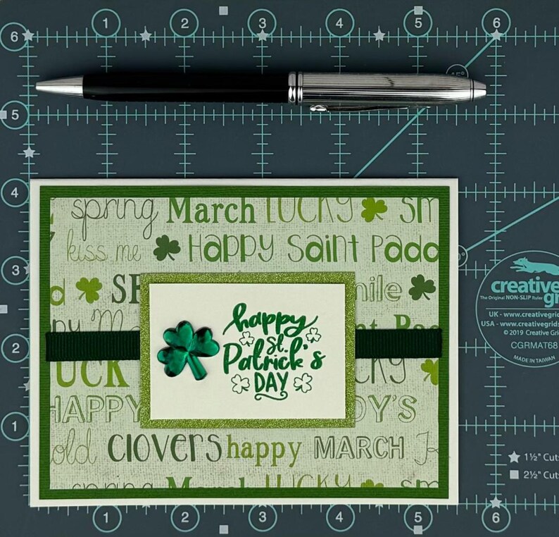 May include: A St. Patrick's Day greeting card with a green and white background featuring the words "Happy St. Patrick's Day" and shamrock designs. A black and silver pen is placed above the card.