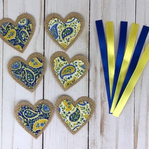 May include: Six heart-shaped burlap patches with paisley fabric centers in blue and yellow. Also, six satin ribbons, alternating between blue and yellow. The items are arranged on a light wood-grain surface.