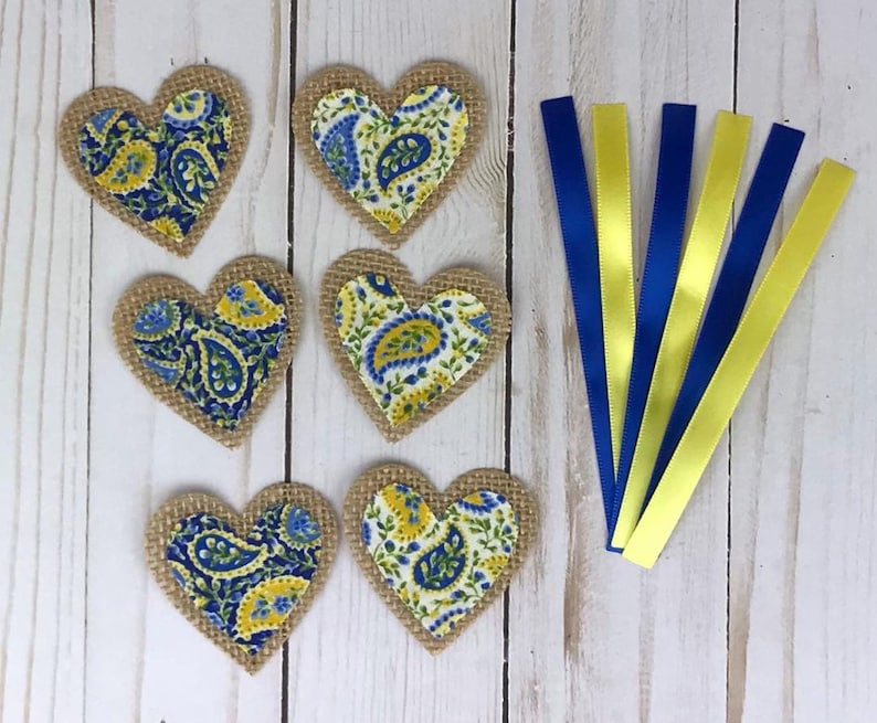 May include: Six heart-shaped burlap patches with paisley fabric centers in blue and yellow. Also, six satin ribbons, alternating between blue and yellow. The items are arranged on a light wood-grain surface.