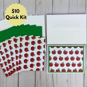 May include: A set of white greeting cards and envelopes featuring a repeating pattern of red apples with green leaves. The cards are arranged in a stack with one card displayed separately to the right, showing a green border around the apple print.