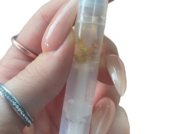 May include: A clear nail cuticle oil pen with a white applicator tip. The pen contains a clear liquid with gold glitter flakes. The pen is held in a hand with long, polished nails and rings.