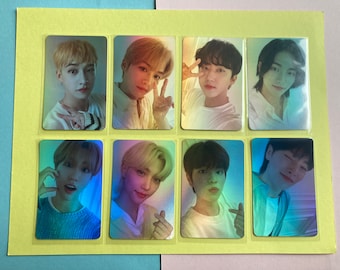 May include: A set of eight holographic photo cards featuring different members of a K-pop group. The cards are arranged in two rows of four, with each card featuring a different member. The cards are all in a similar style, with a holographic background and a close-up photo of the member.