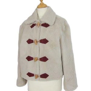 Könnte beinhalten: Beige Kunstfelljacke mit breitem Kragen und drei Knebelverschlüssen aus Holz. Die Jacke hat dunkelrote rautenförmige Akzente um die Knebel. Die Jacke wird auf einer Schaufensterpuppe präsentiert.