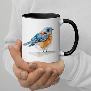 May include: A white ceramic mug with a black rim and handle features a watercolor painting of a bluebird. The bird has a blue head, back, and wings, a brown breast, and a black eye.
