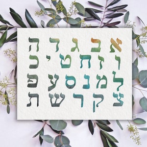 Può includere: Un cartoncino bianco con lettere ebraiche in tonalità di verde e oro, circondato da foglie di eucalipto verdi su uno sfondo bianco.