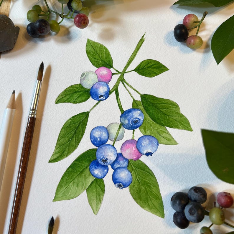May include: A watercolor painting of a blueberry branch with green leaves and clusters of blue, pink, and light-colored berries. The artwork is displayed on white paper alongside a wooden paintbrush and a white pencil, with fresh berries nearby.