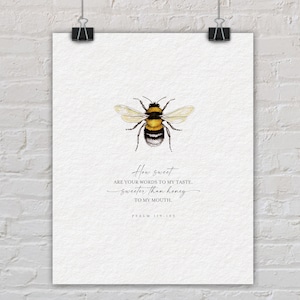 May include: A watercolour print showing a bee in yellow, black, and white. The print includes the text "How sweet are your words to my taste, sweeter than honey to my mouth. Psalm 119:103". The artwork is displayed on a white background and is held by a black clip.