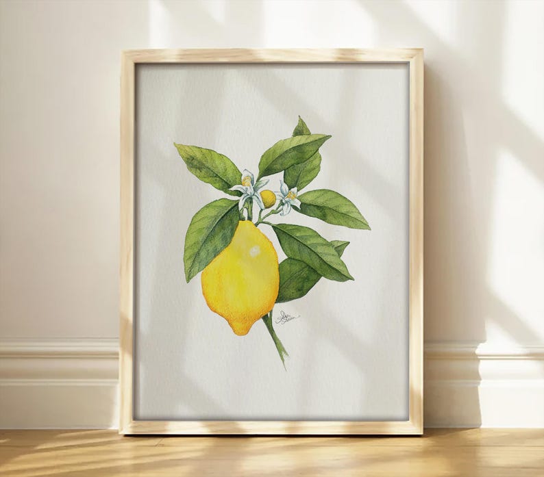 May include: A framed watercolor print featuring a single bright yellow lemon hanging from a green leafy branch with two small white blossoms and a tiny bud. The artwork is centered on a light off-white background within a light wood frame.