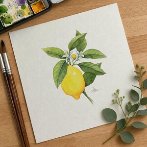 May include: A watercolor painting on white paper depicting a bright yellow lemon hanging from a branch with green leaves and two small white blossoms. The artwork is positioned next to two wooden paintbrushes and a small watercolor paint set.