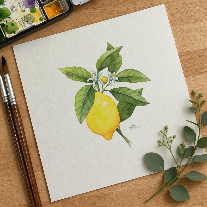 May include: A watercolor painting on white paper depicting a bright yellow lemon hanging from a branch with green leaves and two small white blossoms. The artwork is positioned next to two wooden paintbrushes and a small watercolor paint set.