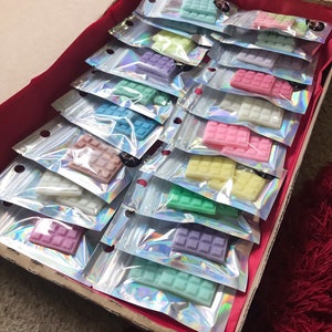 May include: A box filled with small, individually packaged wax melts. The wax melts are in various pastel colours, including pink, blue, green, and purple. Each melt is shaped like a waffle.