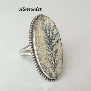 May include: Oval-shaped silver ring featuring a dendritic agate stone. The stone has a cream-colored base with a black, fern-like pattern. The ring has a beaded border and a split shank design. The text "silverindex" is visible.