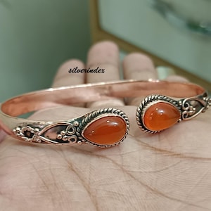 May include: Copper cuff bracelet featuring two teardrop-shaped orange gemstones. The bracelet has an open design with ornate detailing around the gemstones. The metal has a warm, reddish-brown hue, and the overall style is bohemian.