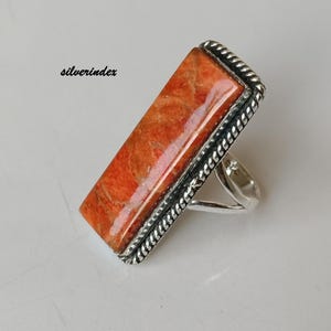May include: A rectangular ring with a vibrant orange stone set in a silver band. The stone has a textured appearance, and the silver band features a detailed, rope-like border. The ring is displayed on a white surface, with the text "silverindex" visible.