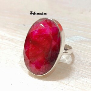 May include: A silver ring with a large, oval-shaped, faceted, red gemstone. The ring is set in a simple, silver band.