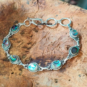 May include: A silver bracelet with a toggle clasp and six oval-shaped abalone shell stones. The bracelet is on a wooden surface. "Silverindex" is printed on the clasp.