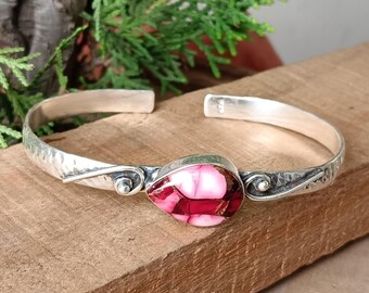 May include: A silver cuff bracelet with a teardrop-shaped, pink and white stone centerpiece. The bracelet has a hammered texture and decorative details near the stone. The stone has a unique, marbled appearance.