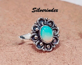 Può includere: Anello d'argento con una pietra preziosa ovale, azzurra e bianca. La pietra è incastonata in una montatura dettagliata a forma di fiore. L'anello è mostrato su uno sfondo neutro, con il testo "Silverindex" visibile.