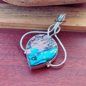 May include: A silver pendant with a teardrop-shaped abalone shell inlay. The shell displays iridescent blues, greens, and purples. The pendant has a decorative silver bail and a loop design. The text "Silverindex" is visible.