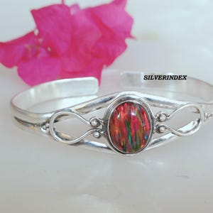 Può includere: Un bracciale a polsino in argento con una gemma ovale multicolore al centro. La gemma mostra sfumature rosse, verdi e gialle. Il bracciale ha un design aperto con anelli decorativi e il testo "SILVERINDEX".
