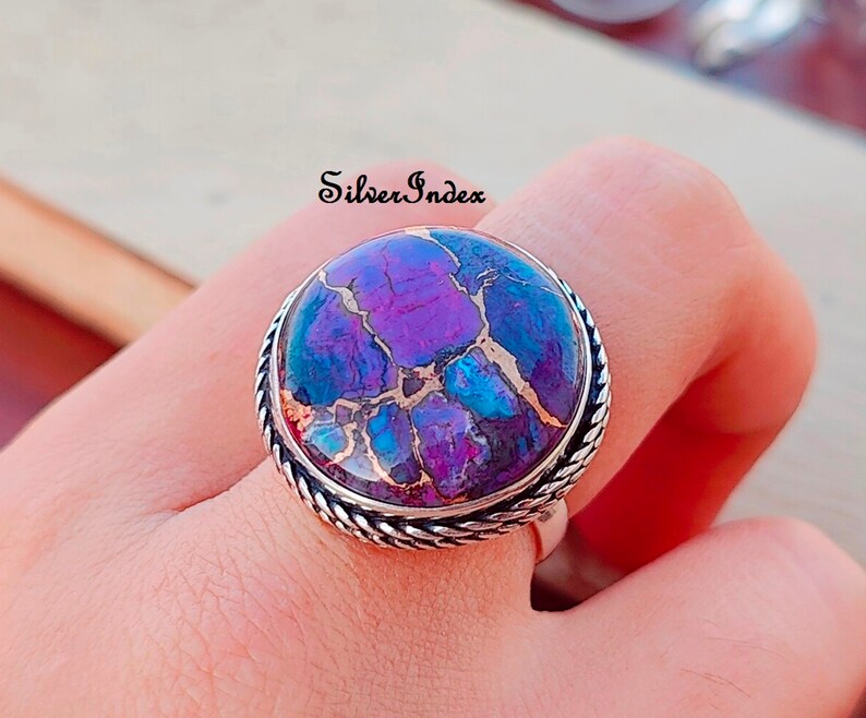 May include: A silver ring with a large, round, purple and blue gemstone set in a silver bezel. The ring has a rope-like design around the bezel.