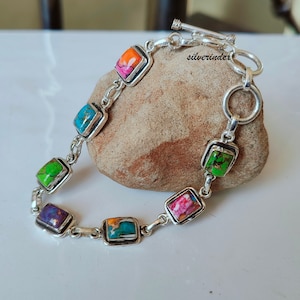 Könnte beinhalten: Ein silbernes Armband mit einem Schiebeverschluss verfügt über sieben quadratische Türkis-Steine in verschiedenen Farben, darunter Grün, Blau, Lila, Rosa und Orange.