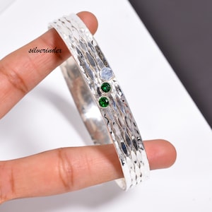Op de afbeelding: Zilveren armband met een gehammerde textuur en drie groene edelstenen. De armband is open en heeft een zilveren index logo.