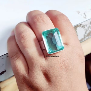 May include: A silver ring with a large, light green, emerald-cut gemstone. The ring is on a person's finger.