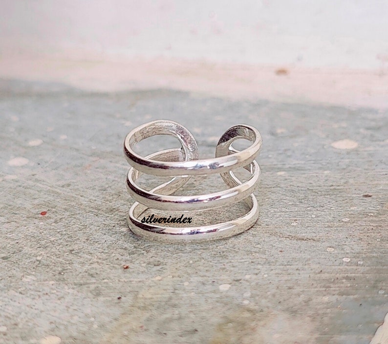 May include: A silver-colored ring with a unique design. The ring features three horizontal bands connected by a vertical loop. The word "silverindex" is engraved on the ring. The ring is made of a shiny, metallic material.