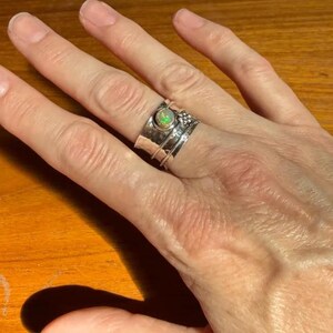 K&ouml;nnte beinhalten: Ein breiter Silberring mit Drehfunktion und einem zentral gefassten, runden Opal, der irisierende gr&uuml;ne und orangefarbene Reflexe zeigt. Die Ringschiene ist geh&auml;mmert und mit einer kleinen dekorativen Ansammlung von Silberkugeln versehen.