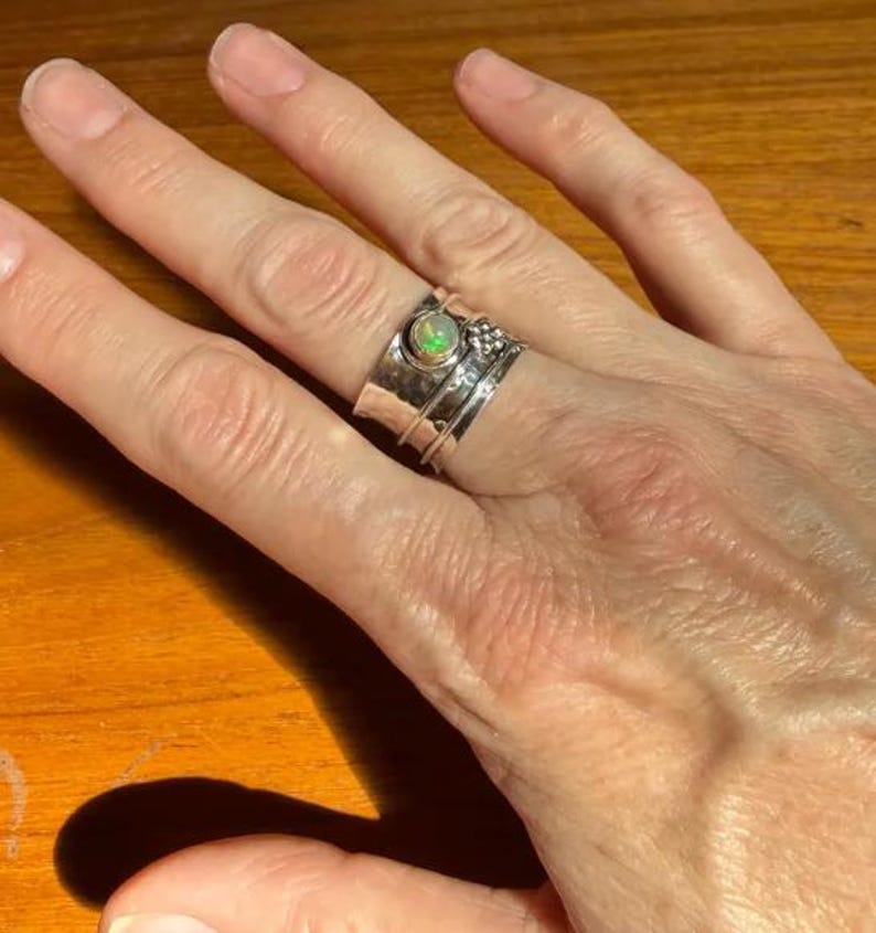 K&ouml;nnte beinhalten: Ein breiter Silberring mit Drehfunktion und einem zentral gefassten, runden Opal, der irisierende gr&uuml;ne und orangefarbene Reflexe zeigt. Die Ringschiene ist geh&auml;mmert und mit einer kleinen dekorativen Ansammlung von Silberkugeln versehen.