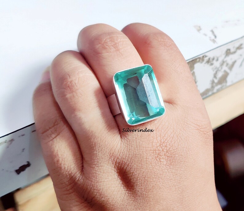 May include: A silver ring with a large, light green, emerald-cut gemstone. The ring is on a person's finger.