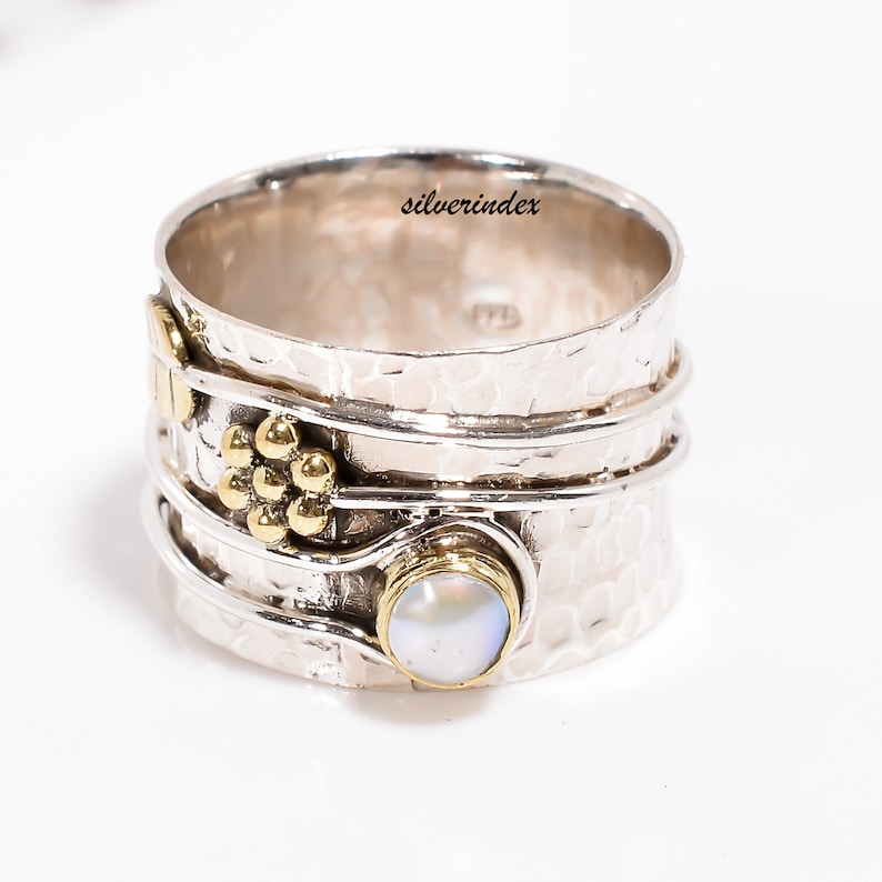 K&ouml;nnte beinhalten: Ein breiter Silberring mit Drehfunktion, geh&auml;mmertem Design, einem goldenen Blumenakzent und einem runden, schillernden Stein in einer goldenen Fassung. Das Band ist mit feinem Silberdraht umwickelt. Der Text silverindex ist zu sehen.