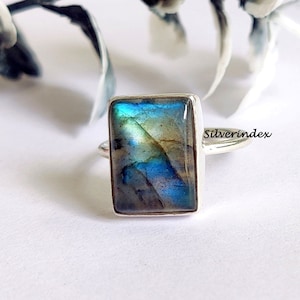 May include: A silver ring featuring a rectangular blue and green labradorite gemstone. The ring is set in a plain band and has a polished finish.