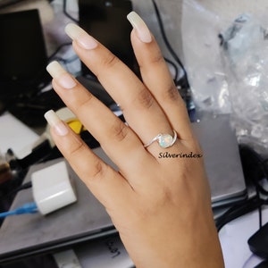 May include: A silver ring with a white opal gemstone. The ring has a curved band and is worn on a hand with long, light-colored nails. The text "Silverindex" is visible in the image.