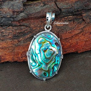 Op de afbeelding: Ovaalvormige hanger met een zilveren frame en een blauwe, groene en iriserende abalone-schelp inlay. De hanger ligt op een houten oppervlak.