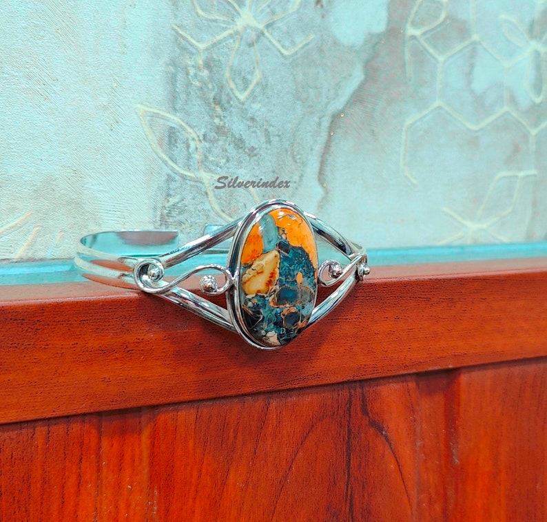 May include: A silver cuff bracelet with an oval-shaped, multi-colored gemstone set in the center. The gemstone has shades of orange, blue, and brown. The bracelet has a delicate, intricate design.