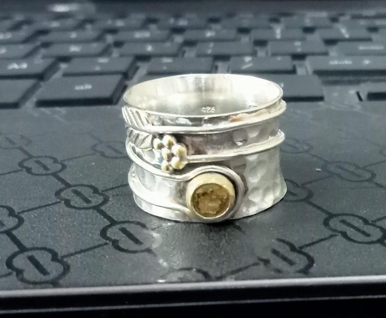 K&ouml;nnte beinhalten: Ein breiter Spinner-Ring aus Sterlingsilber mit geh&auml;mmerter Oberfl&auml;che, einem kleinen goldfarbenen Blumenakzent und einer runden, goldfarbenen Steinfassung. Die Innenseite ist mit 925 gestempelt, was den Silbergehalt dieses Rings kennzeichnet.