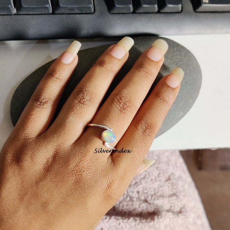 May include: A silver ring with a white opal gemstone. The ring is designed with a curved band and the opal is set in a bezel setting. The ring is worn on a person's finger.