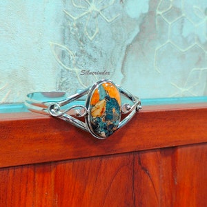 May include: A silver cuff bracelet with an oval-shaped, multi-colored gemstone set in the center. The gemstone has shades of orange, blue, and brown. The bracelet has a delicate, intricate design.