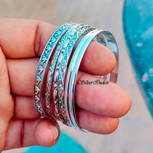 May include: A set of four silver bangles with intricate floral and bird designs. The bangles are stacked on top of each other and are worn on a person's wrist.