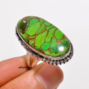May include: A silver ring with a large oval green turquoise stone set in a silver bezel. The stone has a copper matrix and a green and brown color.