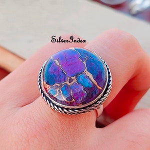 May include: A silver ring with a large, round, purple and blue gemstone set in a silver bezel. The ring has a rope-like design around the bezel.